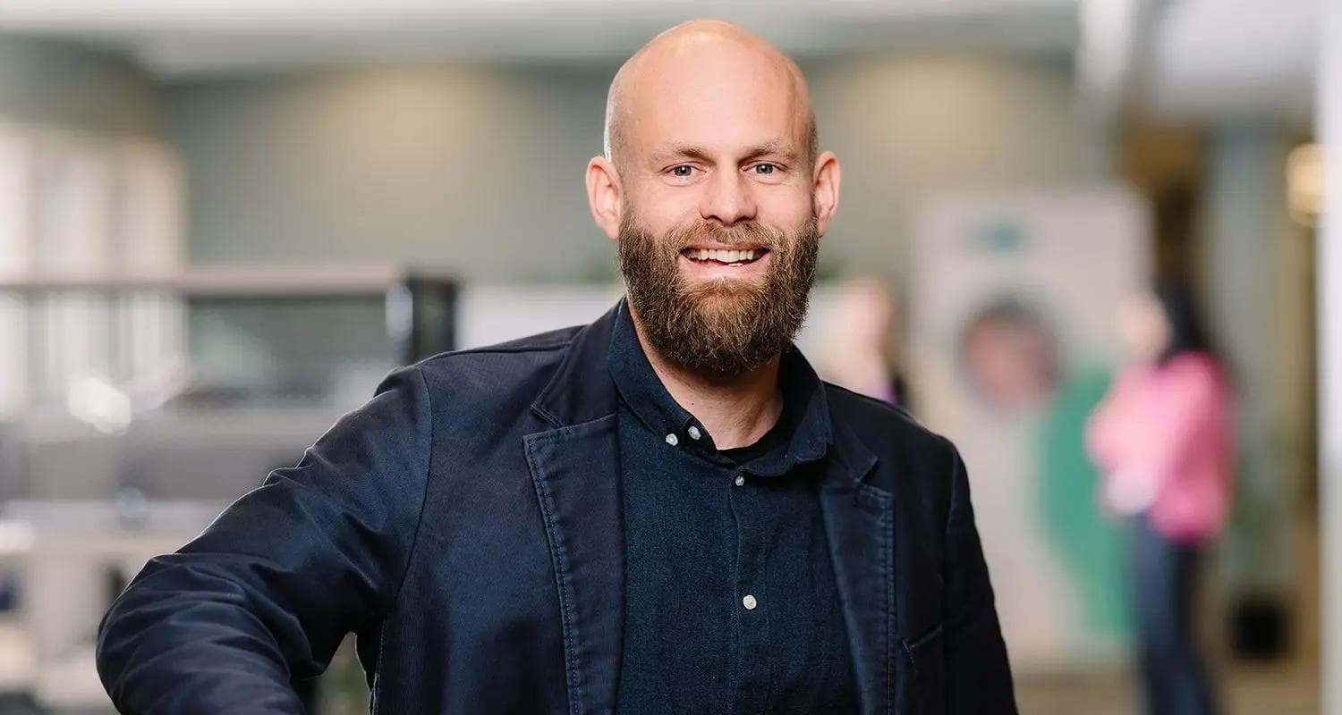 Smilende skallet mann med skjegg og mørk blazer står i et moderne kontormiljø med uskarpe bakgrunnsdetaljer.