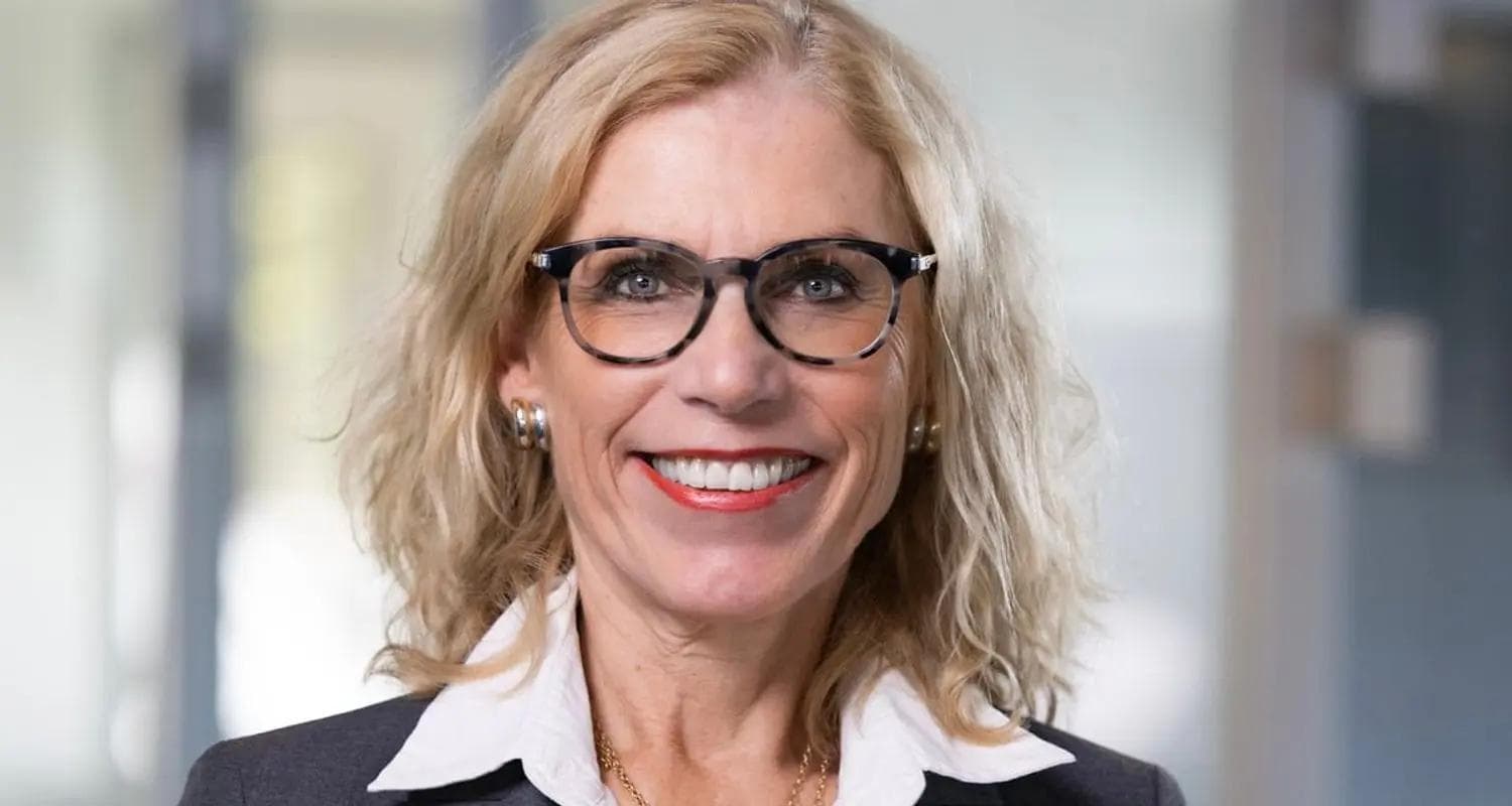 milende kvinne med blondt hår, briller og dressjakke med hvit skjorte står i et moderne kontormiljø med glassvinduer.