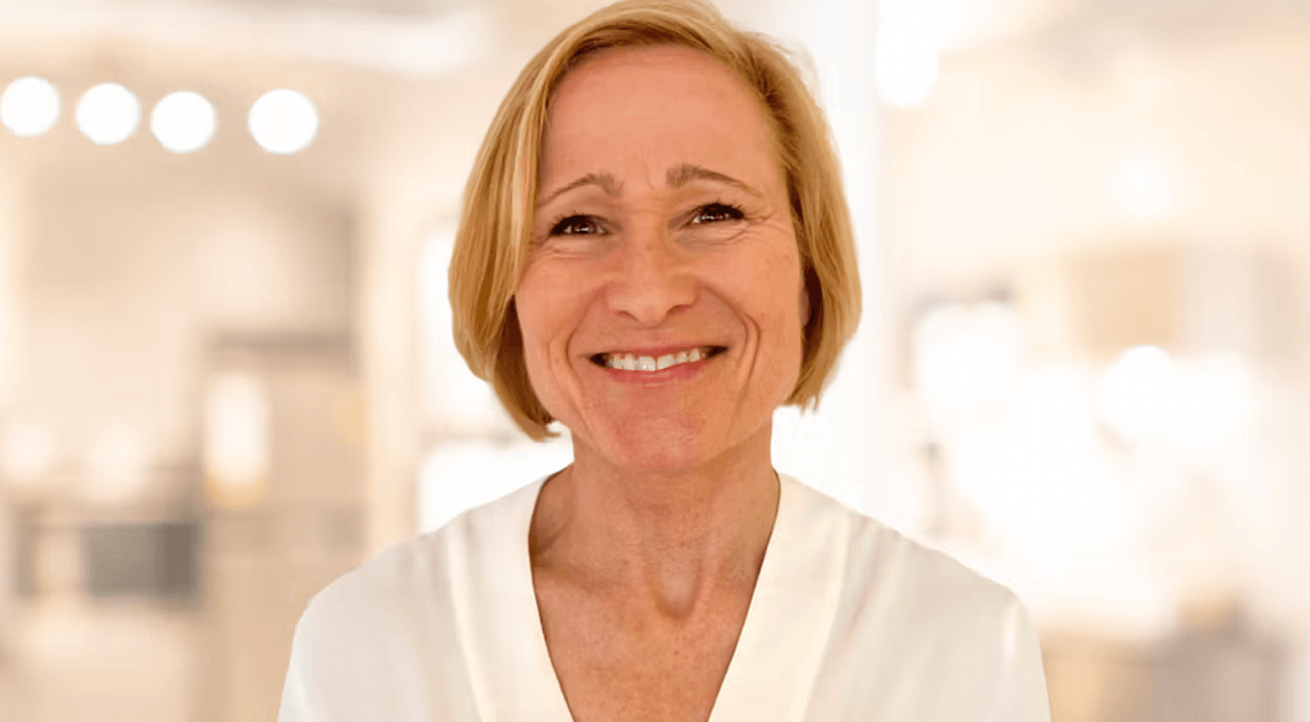 Smilende kvinne med kort blondt hår og hvit bluse står i et lyst, uskarpt innendørsmiljø med varm belysning.