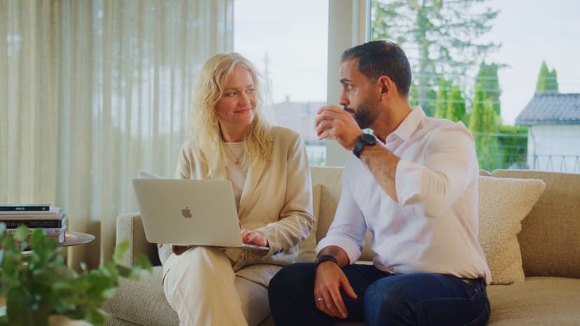 Kvinne med laptop og mann i hvit skjorte sitter i en lys stue. De smiler og diskuterer, med grønn utsikt gjennom vinduet.