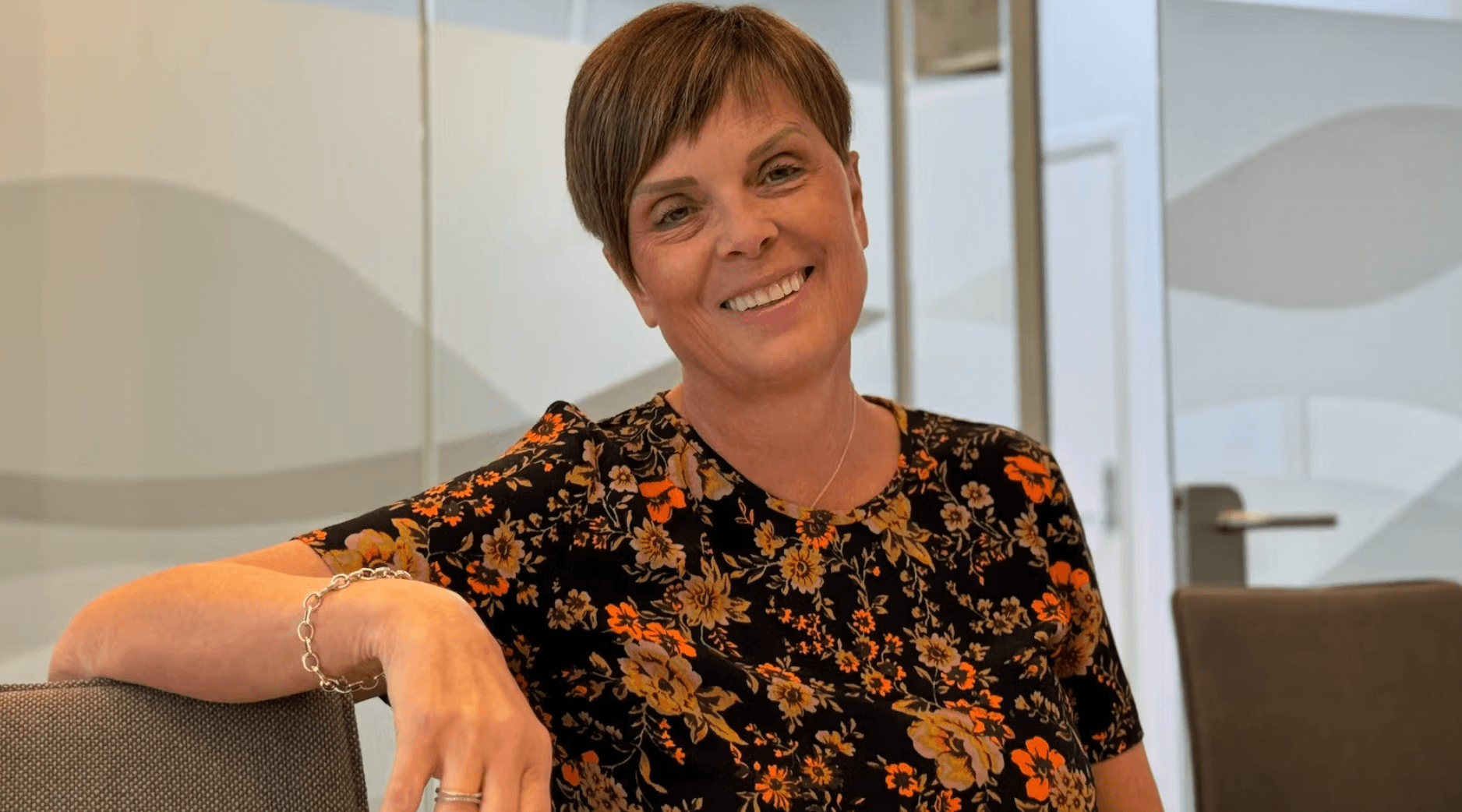 Smilende kvinne med kort mørkt hår og blomstermønstret bluse sitter avslappet i et moderne kontormiljø med glassvegger.
