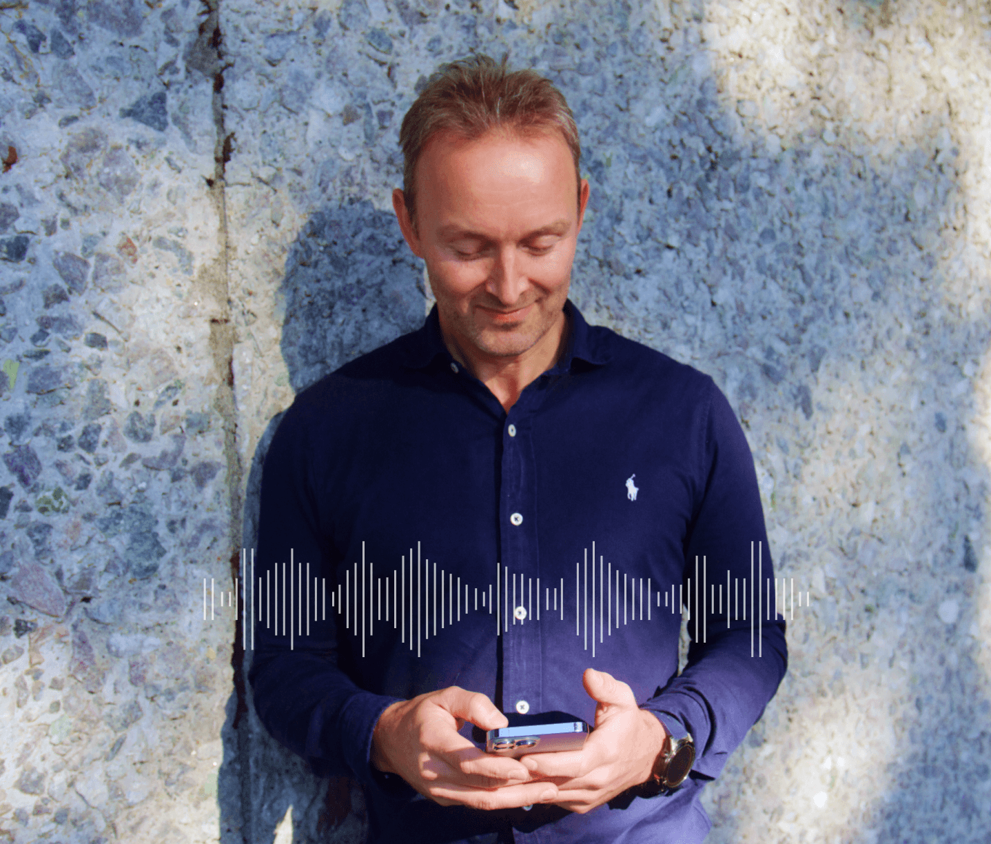 Smilende mann i mørk blå skjorte står mot en steinvegg og ser på mobilen. Lydbølgesymbol vises over skjermen.